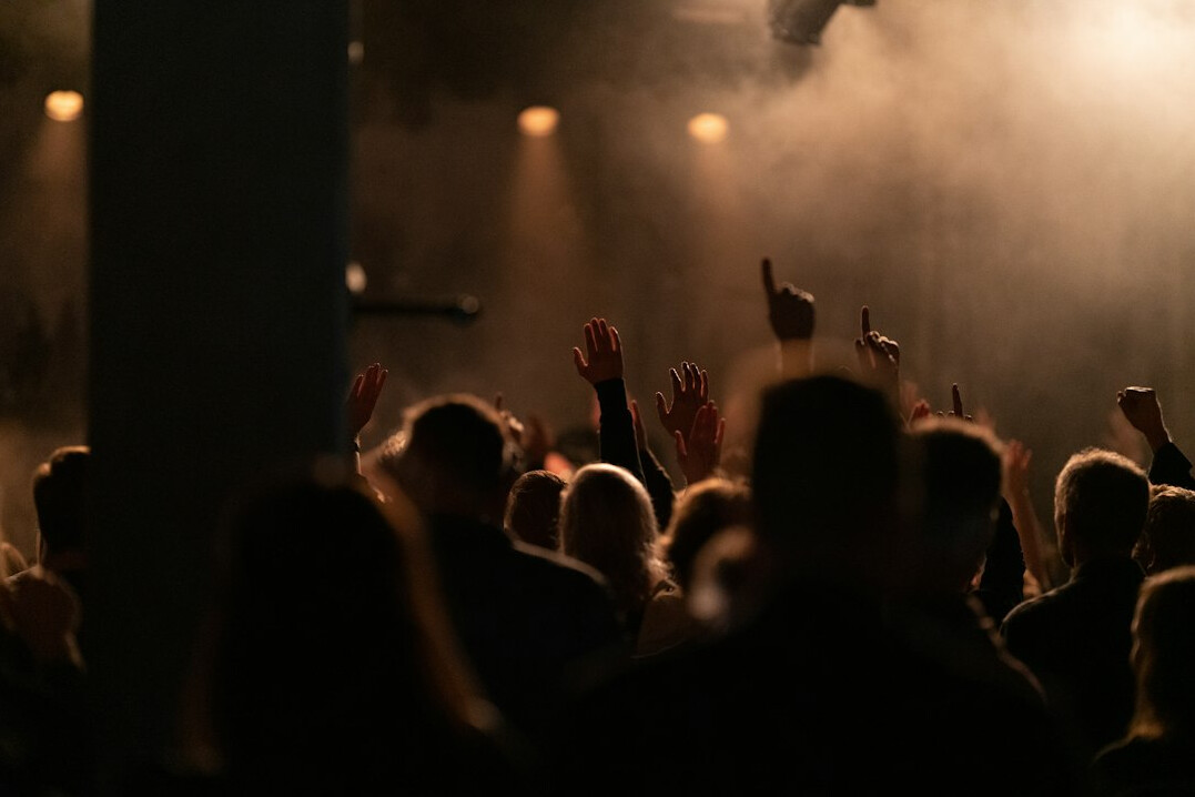 Crowd of audience members seen from behind facing a softly lit concert stage